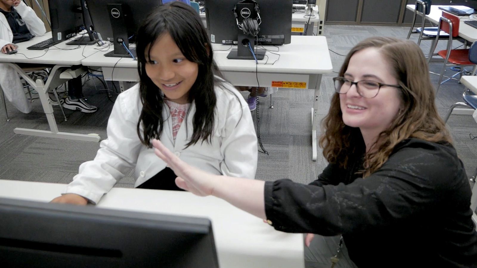 A teacher crouches beside a student at a computer, and both are smiling.