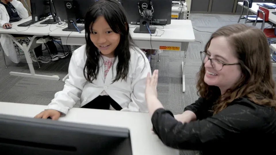 A smiling teacher sits beside a student in a lab coat at her computer, raising a hand toward her for a high five.