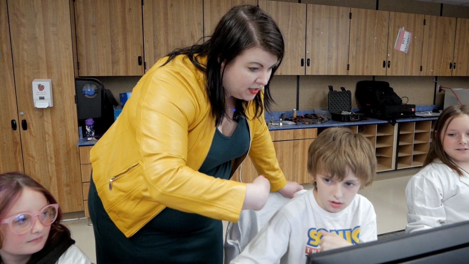 A teacher in a yellow jacket bends down to talk a student through the next step.