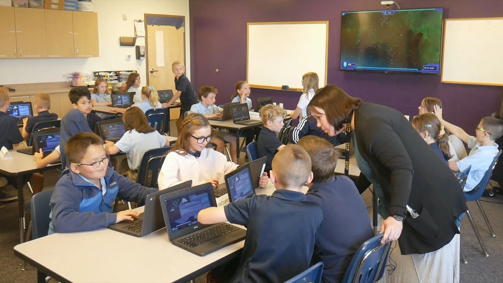 Upper-elementary students work at rows of Chromebooks while a teacher in a black cardigan leans down to two boys sharing a table, and a dark Mission screen glows on the wall behind them.