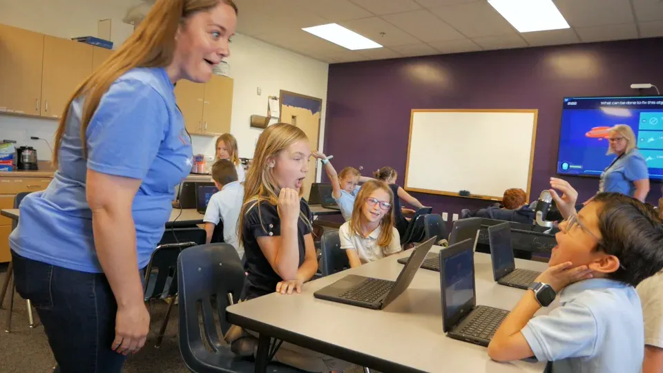 A teacher leans in with a grin as her students react to the Mission on the screen.