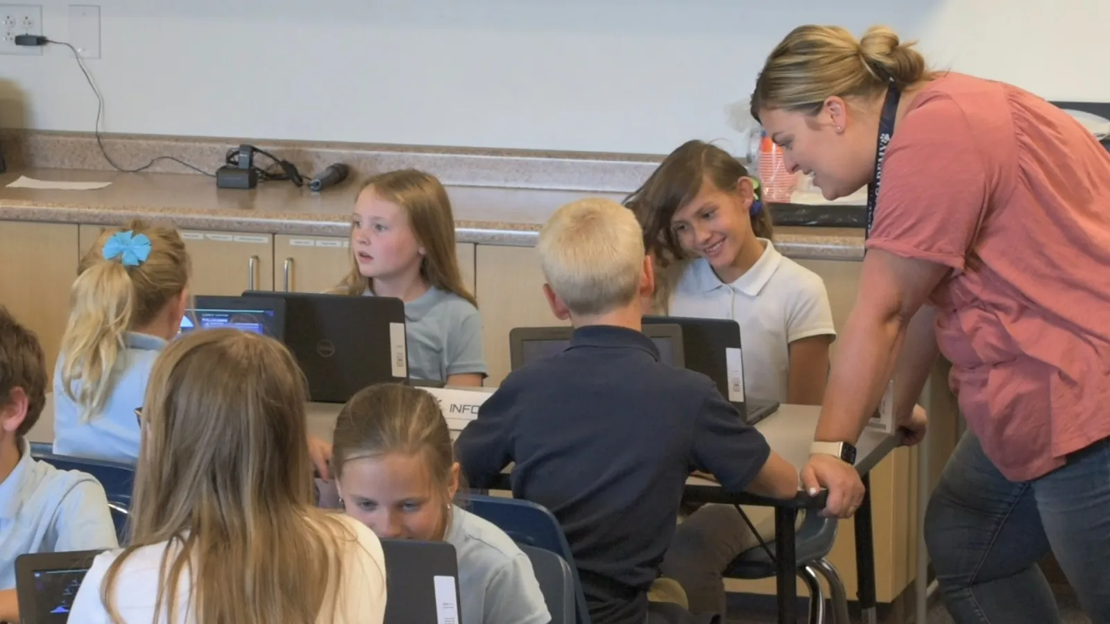 A teacher leans in to an upper-elementary team at their Chromebooks, and two students look up at her, grinning.