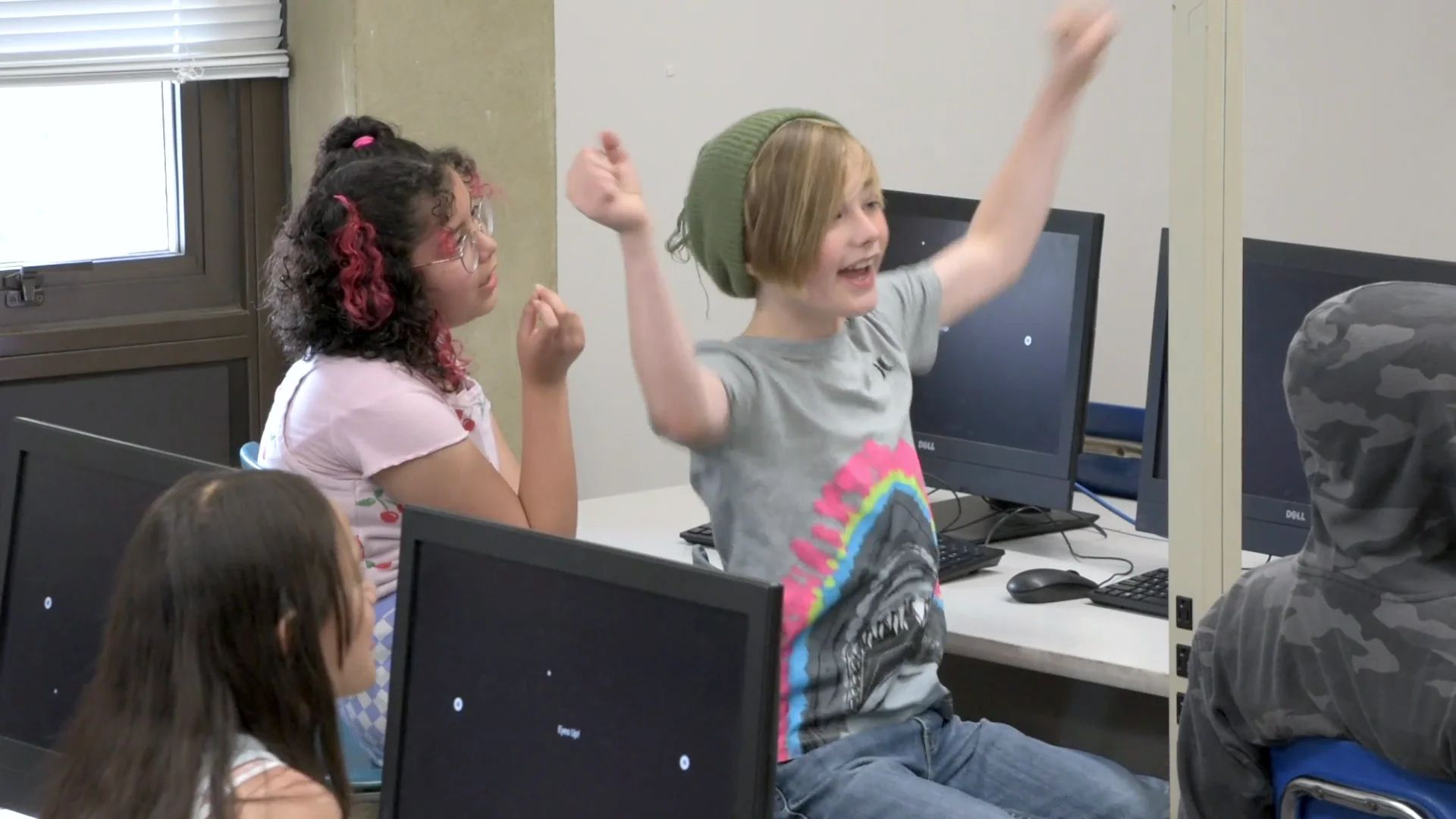 A student in a green beanie throws both arms up mid-cheer at a computer lab desk.