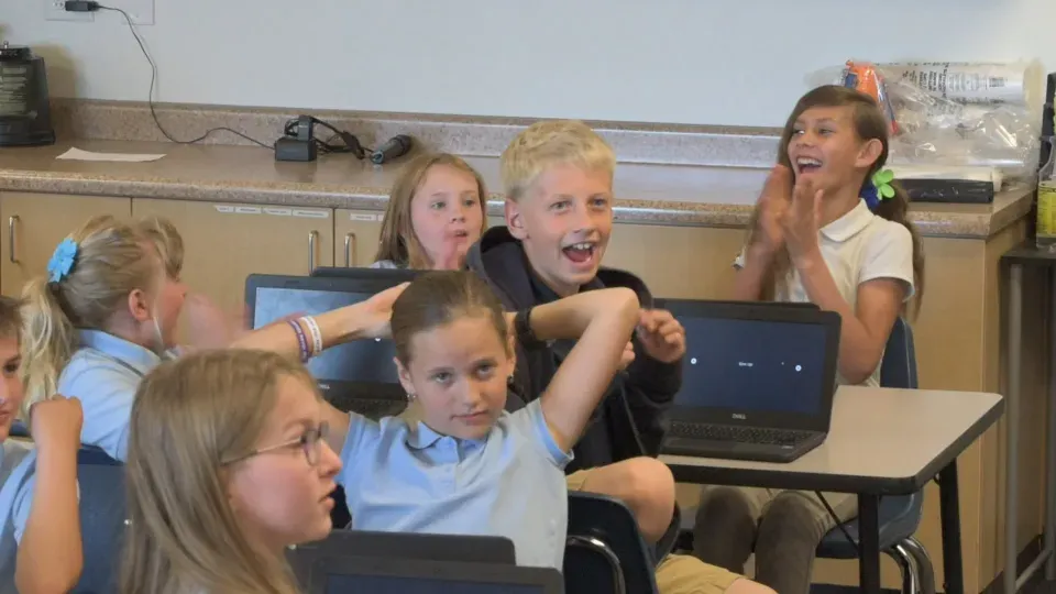 Young students in school uniforms cheer and clap at their laptops, one girl with both hands on top of her head.