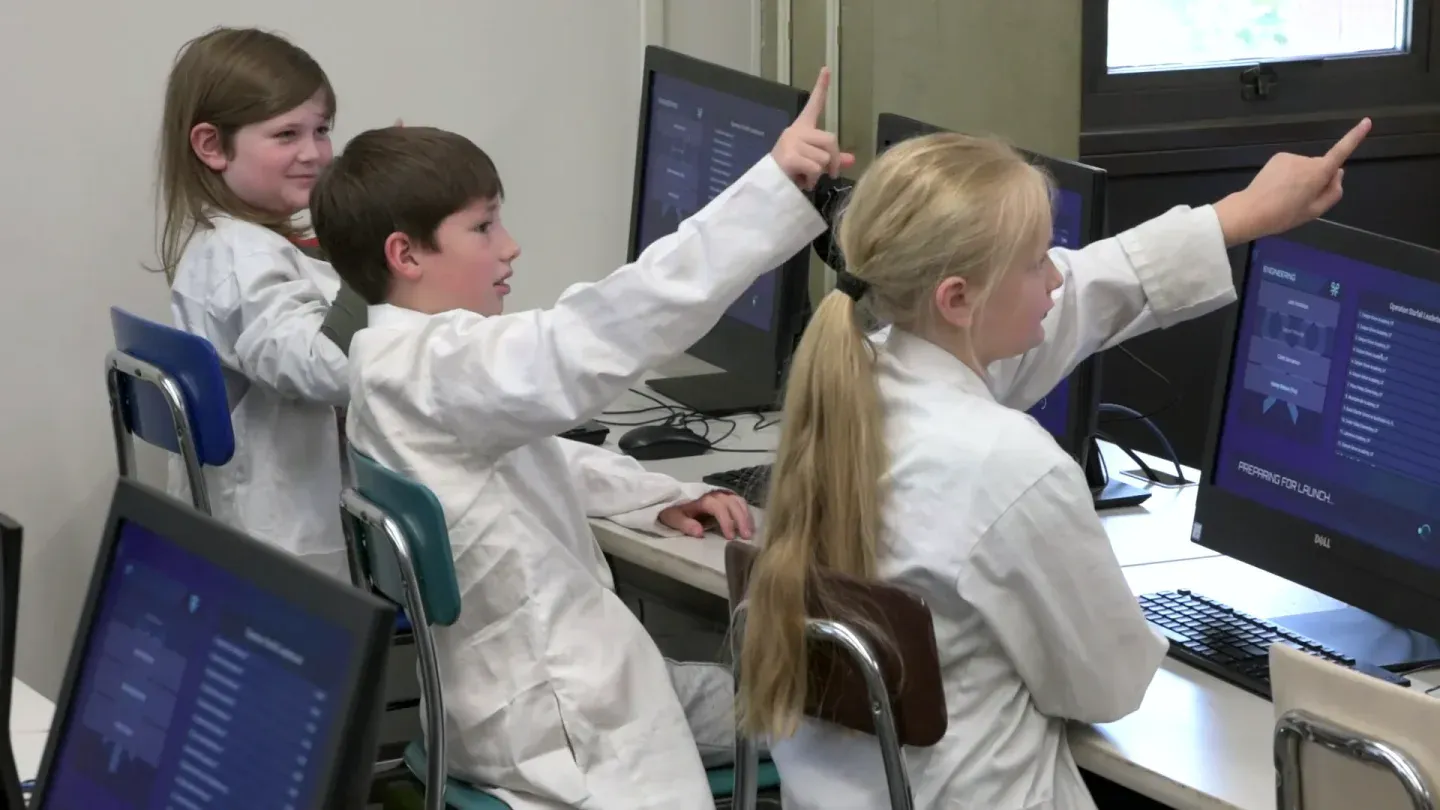 Three students in white lab coats point toward the front of the room from their computer stations; one screen reads Preparing for launch.