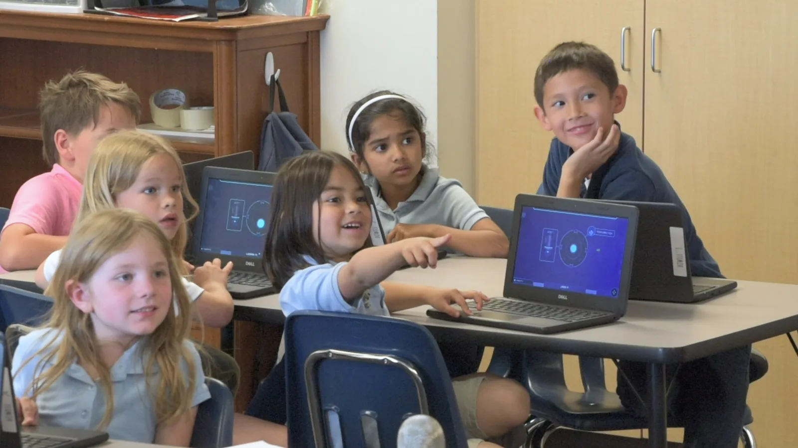 Young students look up from their Chromebooks, one pointing toward the front of the room as a classmate grins.