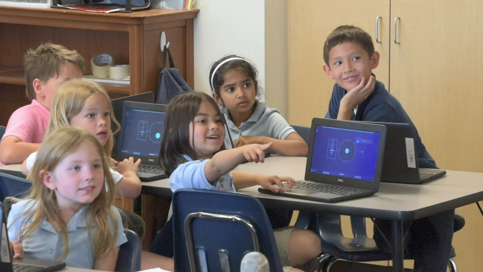 Young students look up from their Chromebooks, one pointing toward the front of the room as a classmate grins.