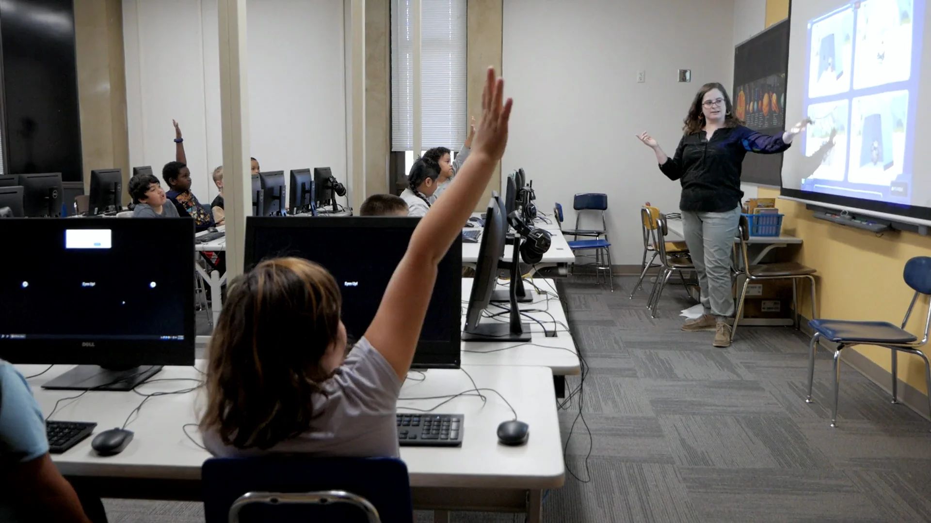 A teacher at the front of a computer lab gestures at the Mission on the screen as a student in the foreground stretches a hand high.