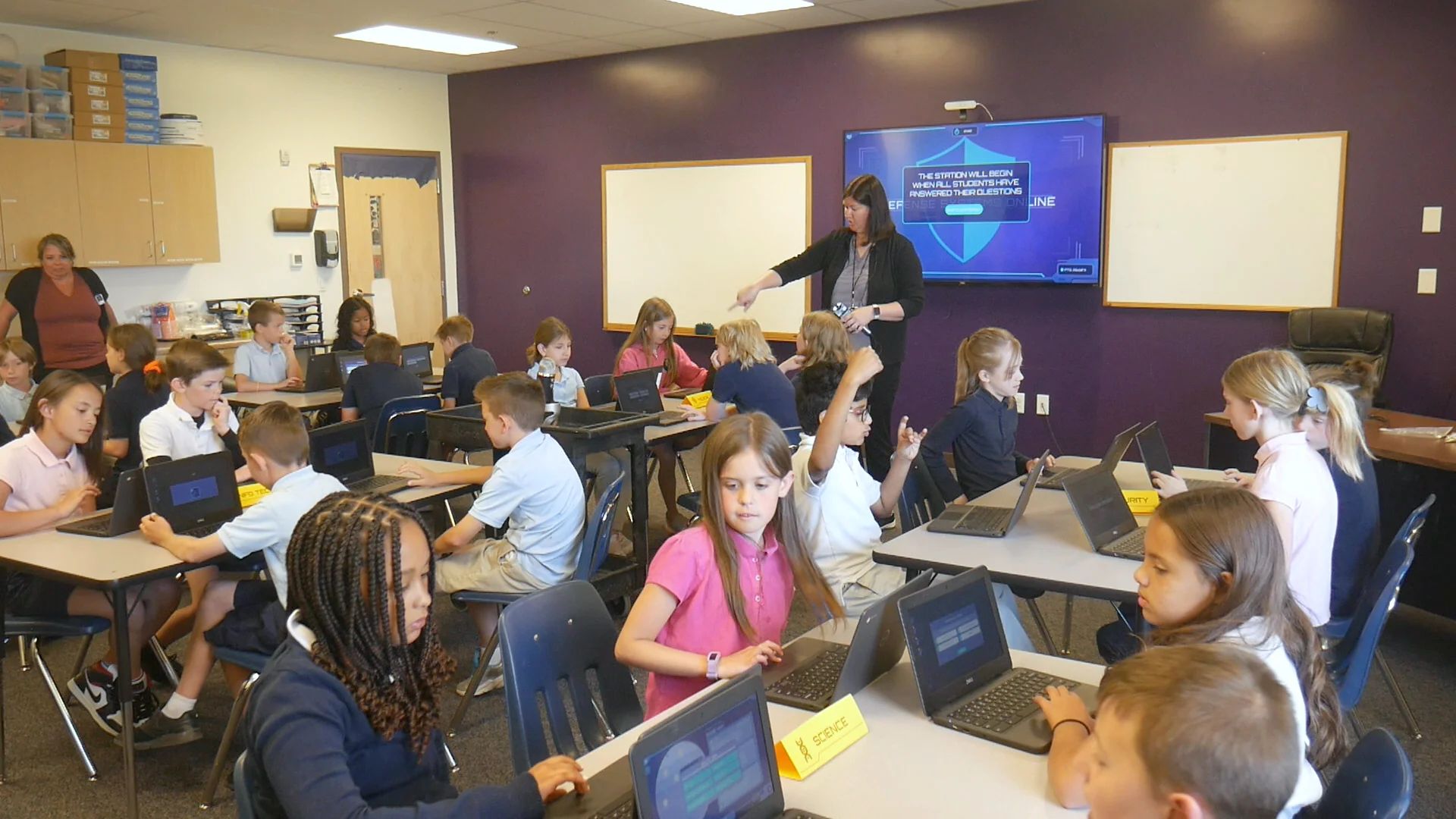 A teacher points at the Mission on the wall screen while her class works at their Chromebooks, a boy raising his hand.