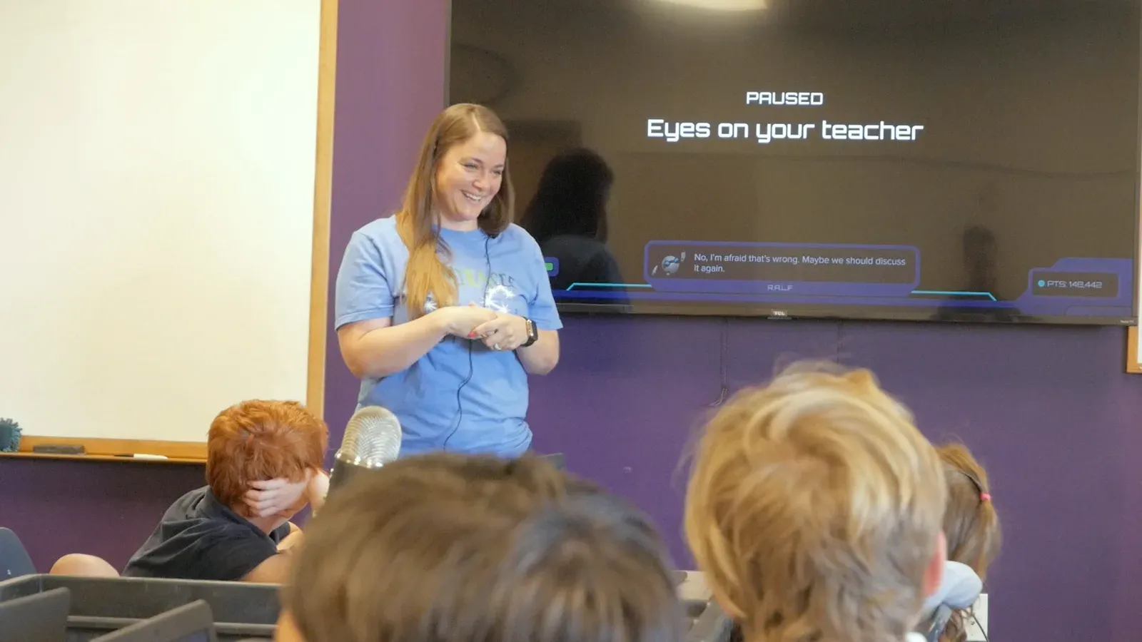 A teacher laughs at the front of her class while the Mission is paused on the screen behind her: Eyes on your teacher.