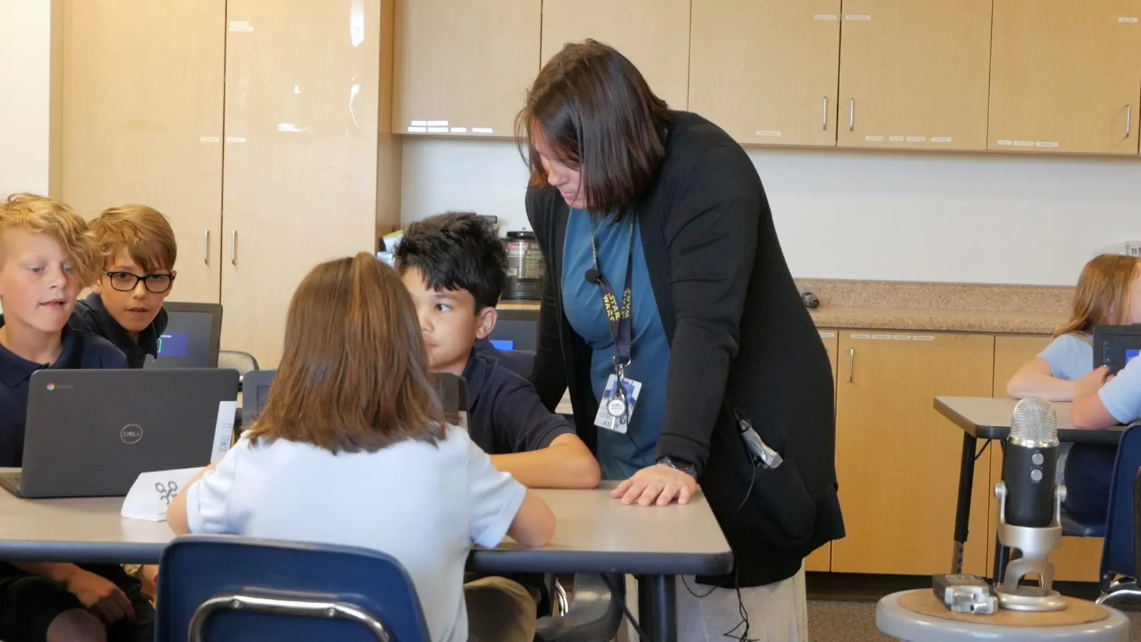 A teacher in a black cardigan leans over a table of students at their Chromebooks, while two boys at the end of the table look across at their teammates.