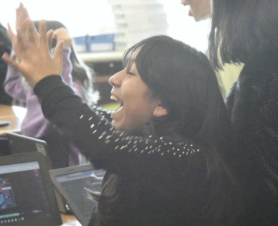 A student throws both hands up and cheers mid-Mission, her Chromebook open in front of her.