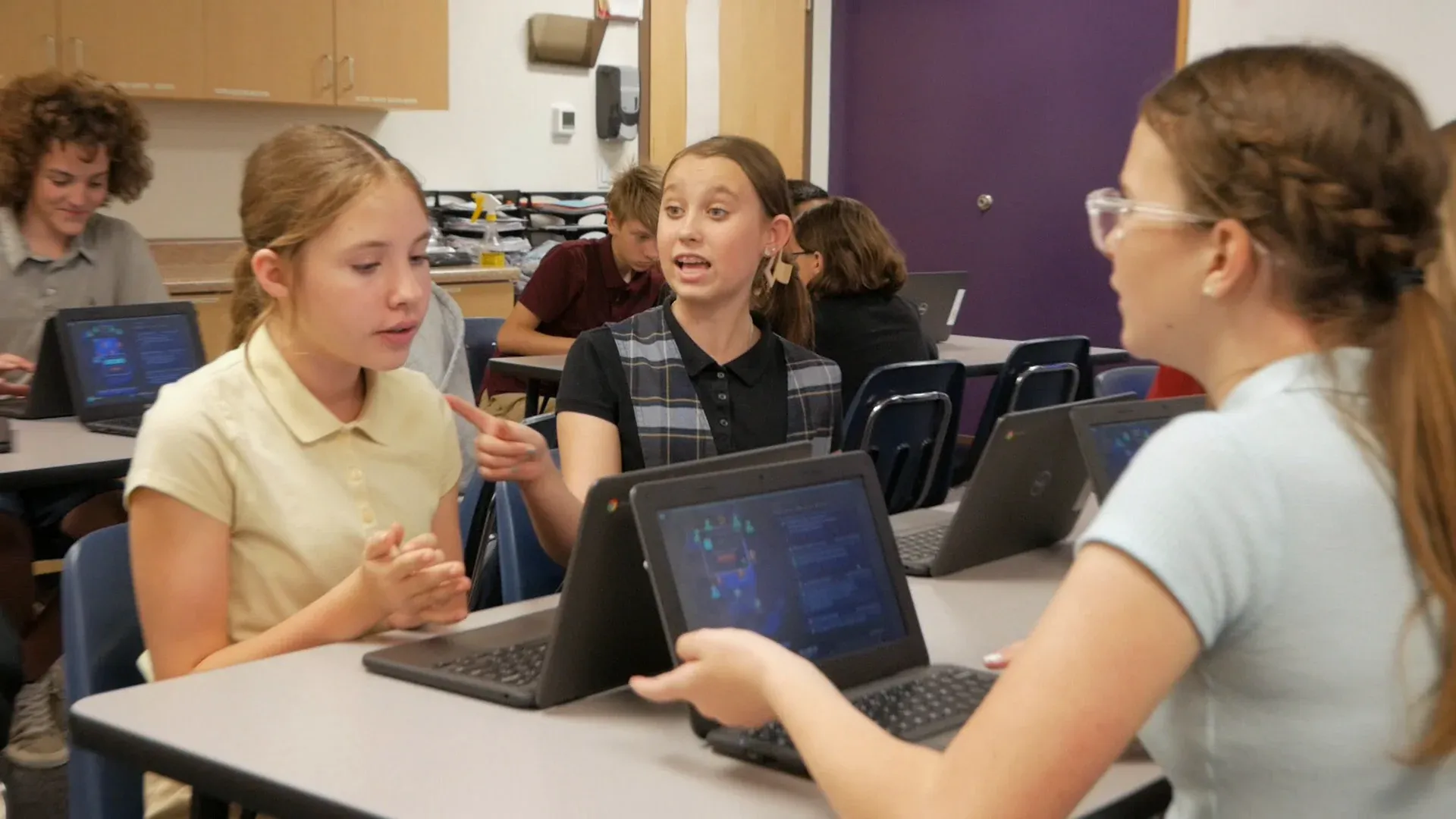 Three middle schoolers at their Chromebooks argue a point across the desks, one pointing as she makes it.