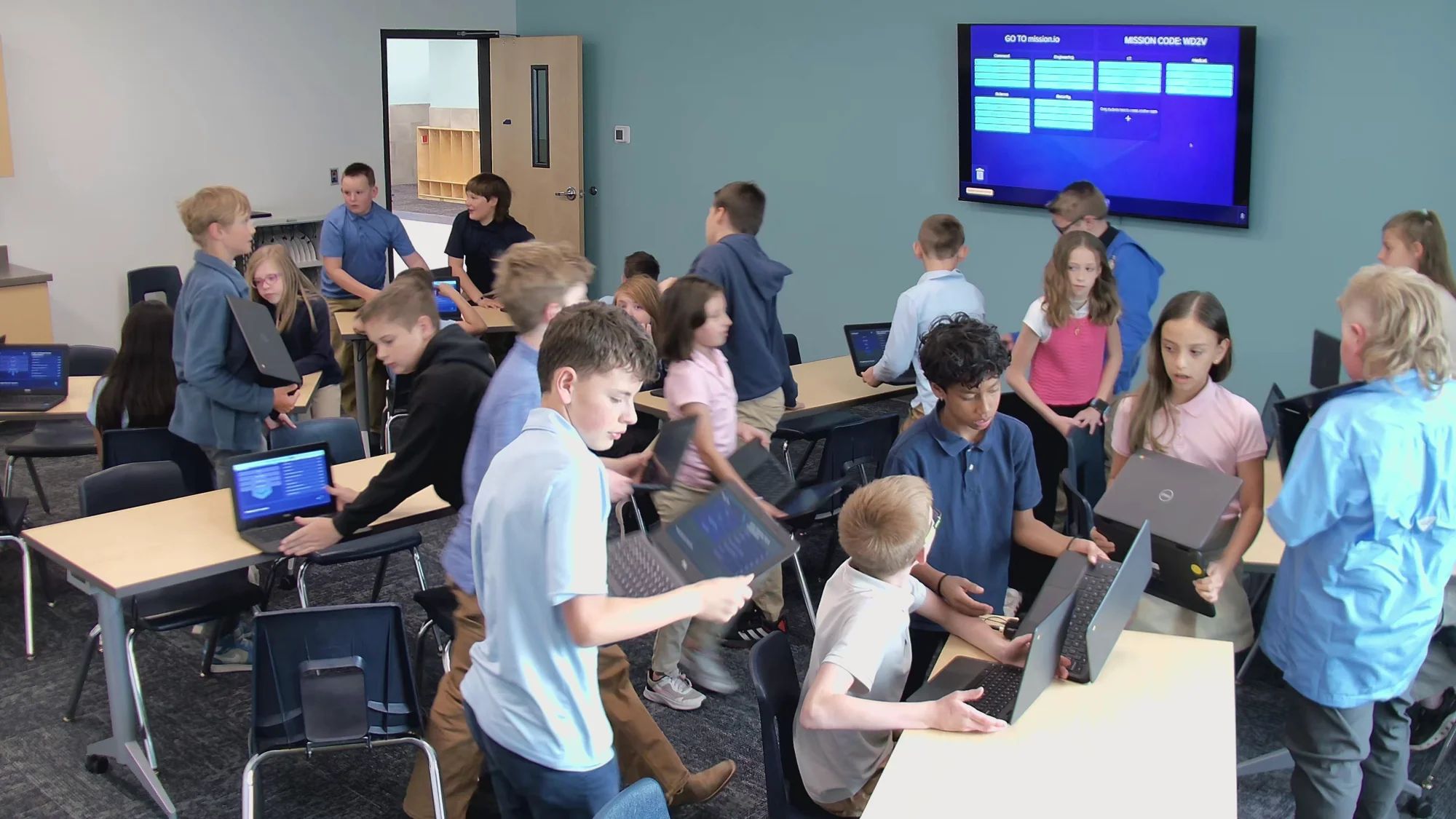 Teams of students gather around their desks in a busy classroom, the Mission on the wall screen behind them.