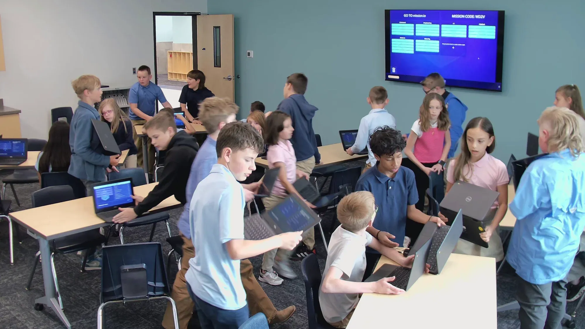 An upper-elementary class moves into crews with their Chromebooks while the screen at the front lists the stations.