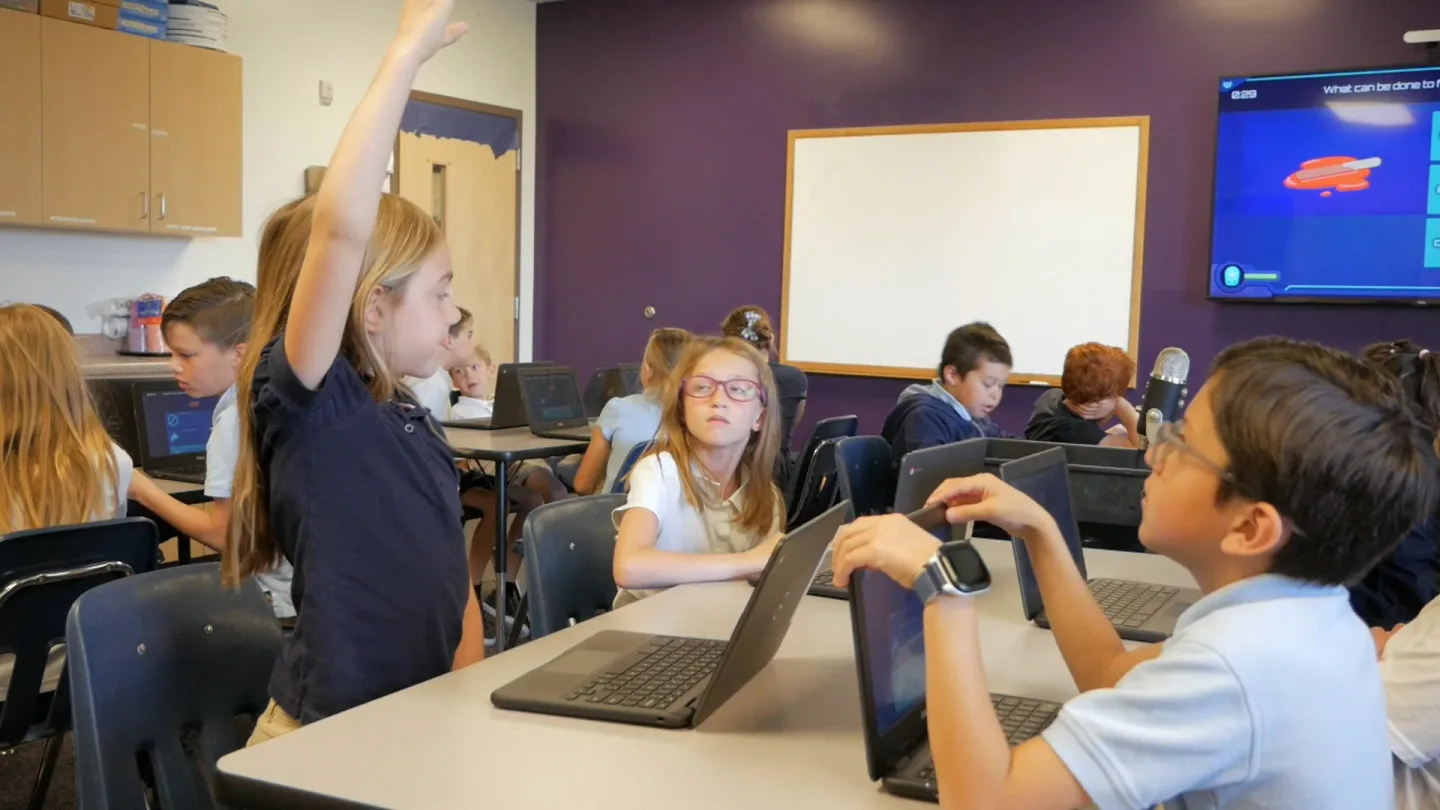 A student stretches her hand high while classmates look up at her and the question on the wall screen.