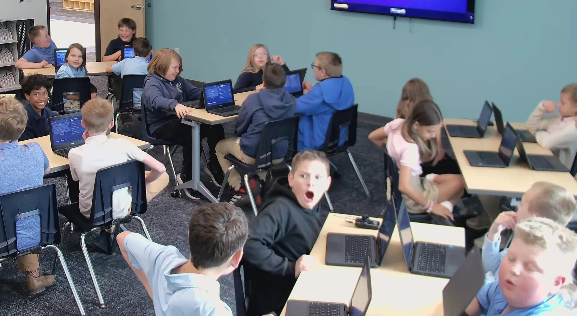 A boy turns with an open-mouthed gasp while classmates work and laugh at the tables behind him.