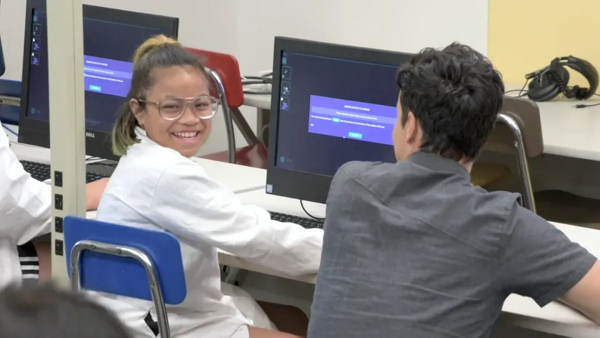 A student in a lab coat and goggles grins over her shoulder from her computer during a Mission.