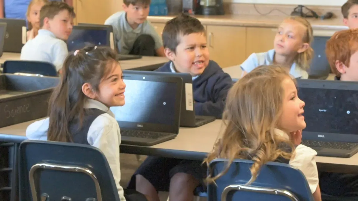 Young students at their Chromebooks turn to each other, laughing, in the middle of a Mission.