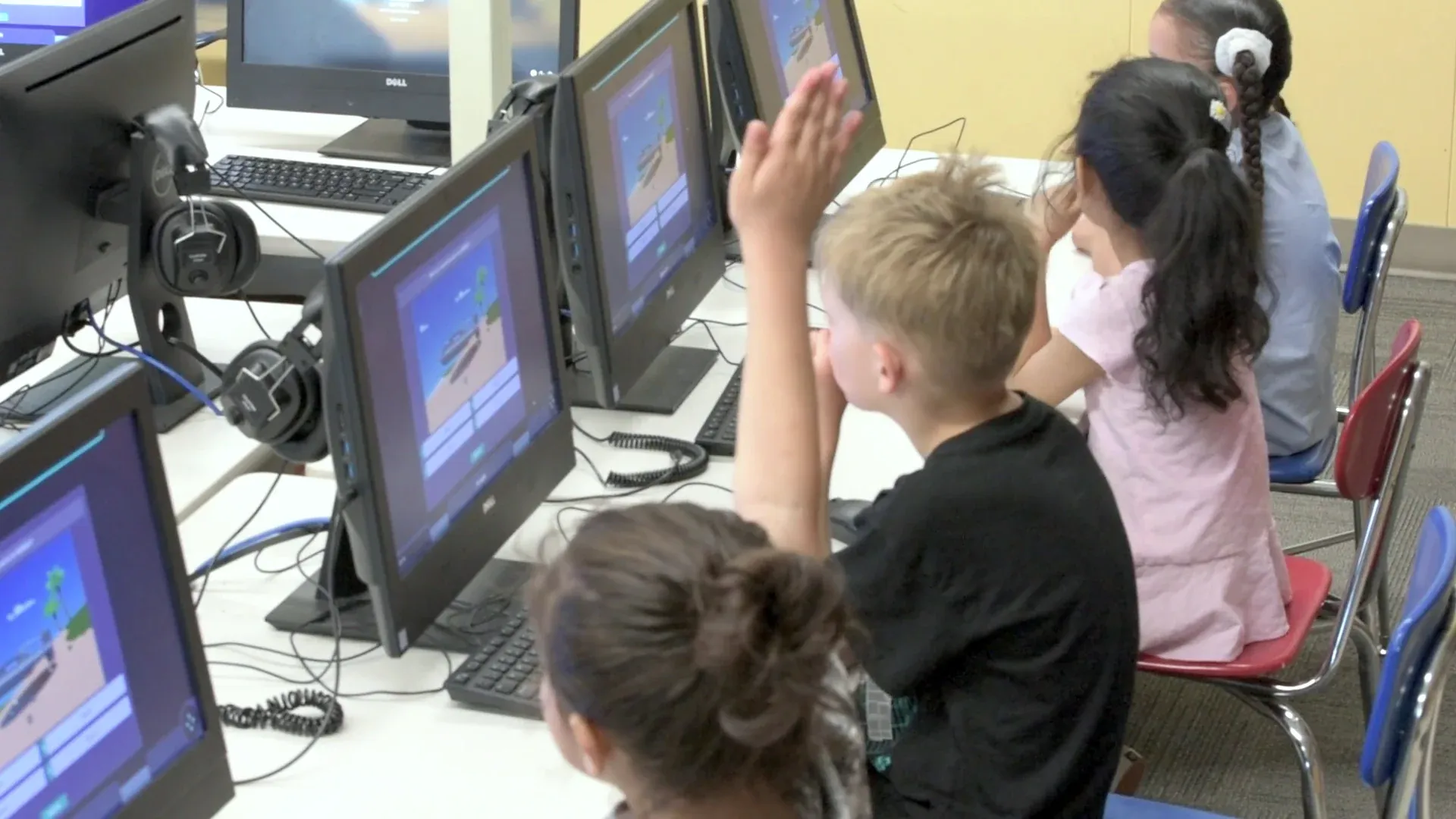 A young boy shoots his arm up to answer beside a classmate, rows of monitors showing the Mission.
