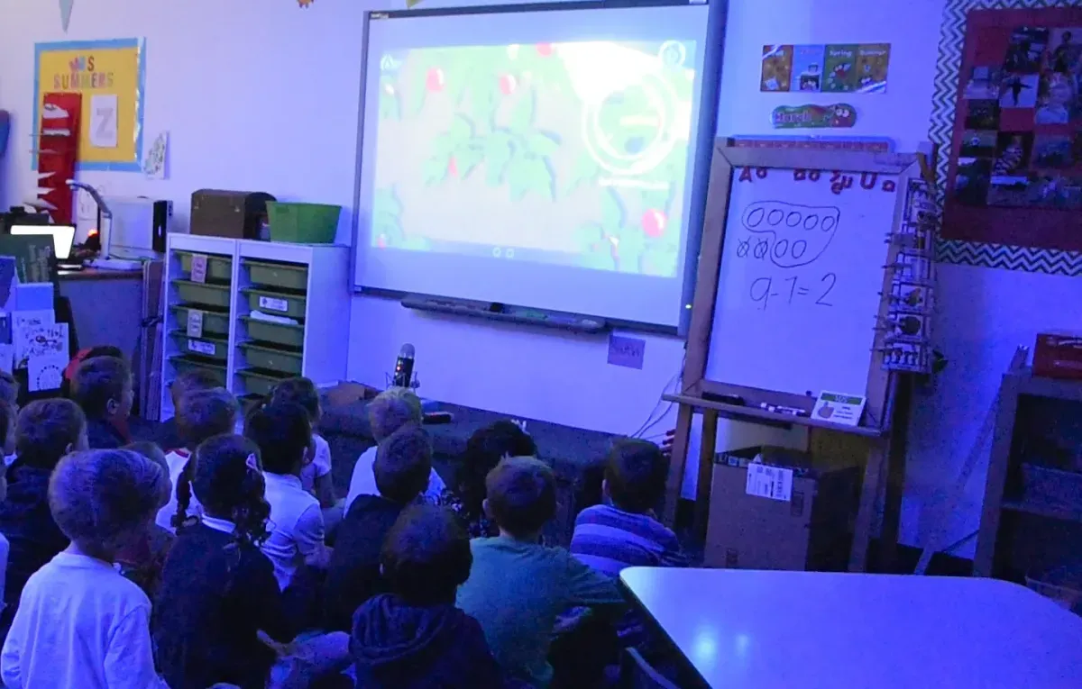 A class sits on the carpet in a dim room, every face turned up to the Mission on the projector screen.
