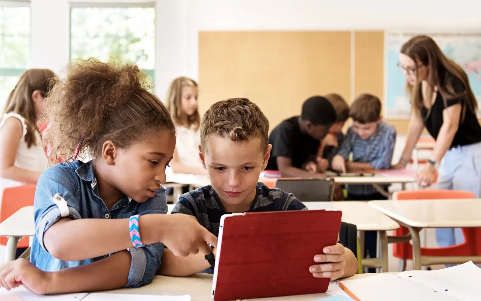 Two elementary students share a red tablet at their desk while a teacher helps classmates working together in the background.