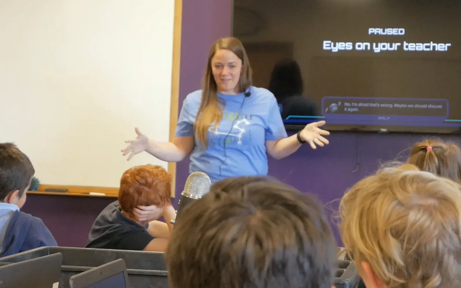 A teacher talks with her class while the board behind her reads Paused, Eyes on your teacher.