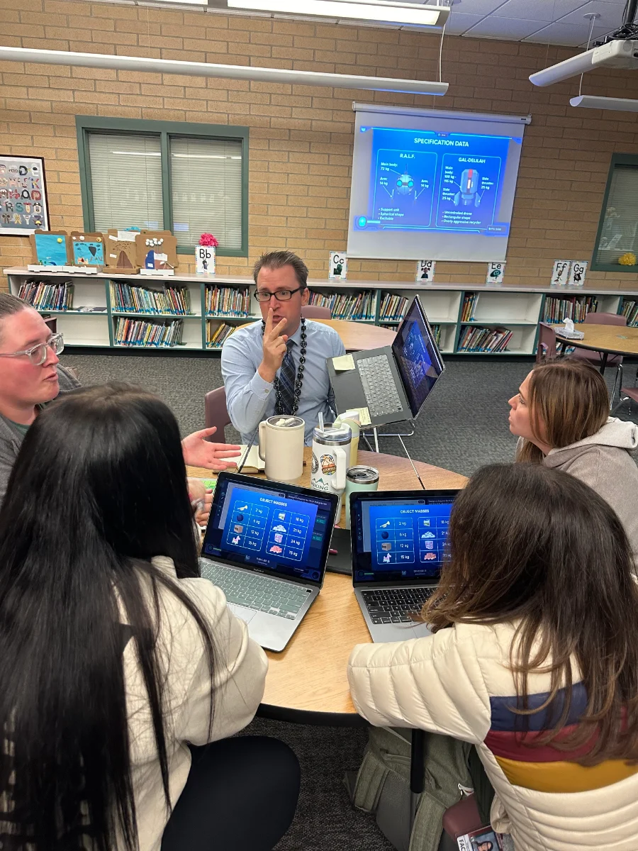 A teacher mid-sentence at a library table while colleagues work the Mission on their laptops.