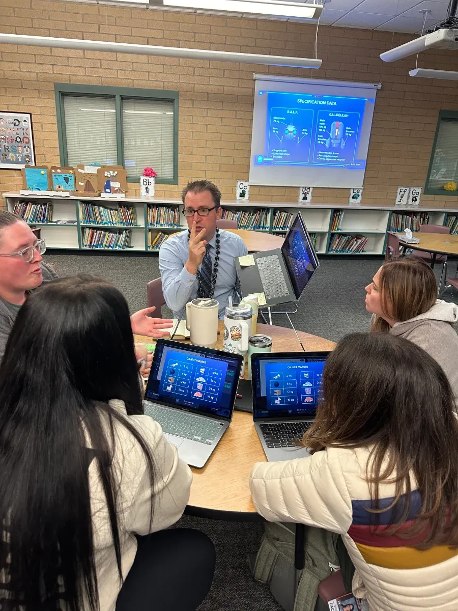Five teachers around a library table work a Mission on their laptops while one, chin in hand, thinks through the next call.