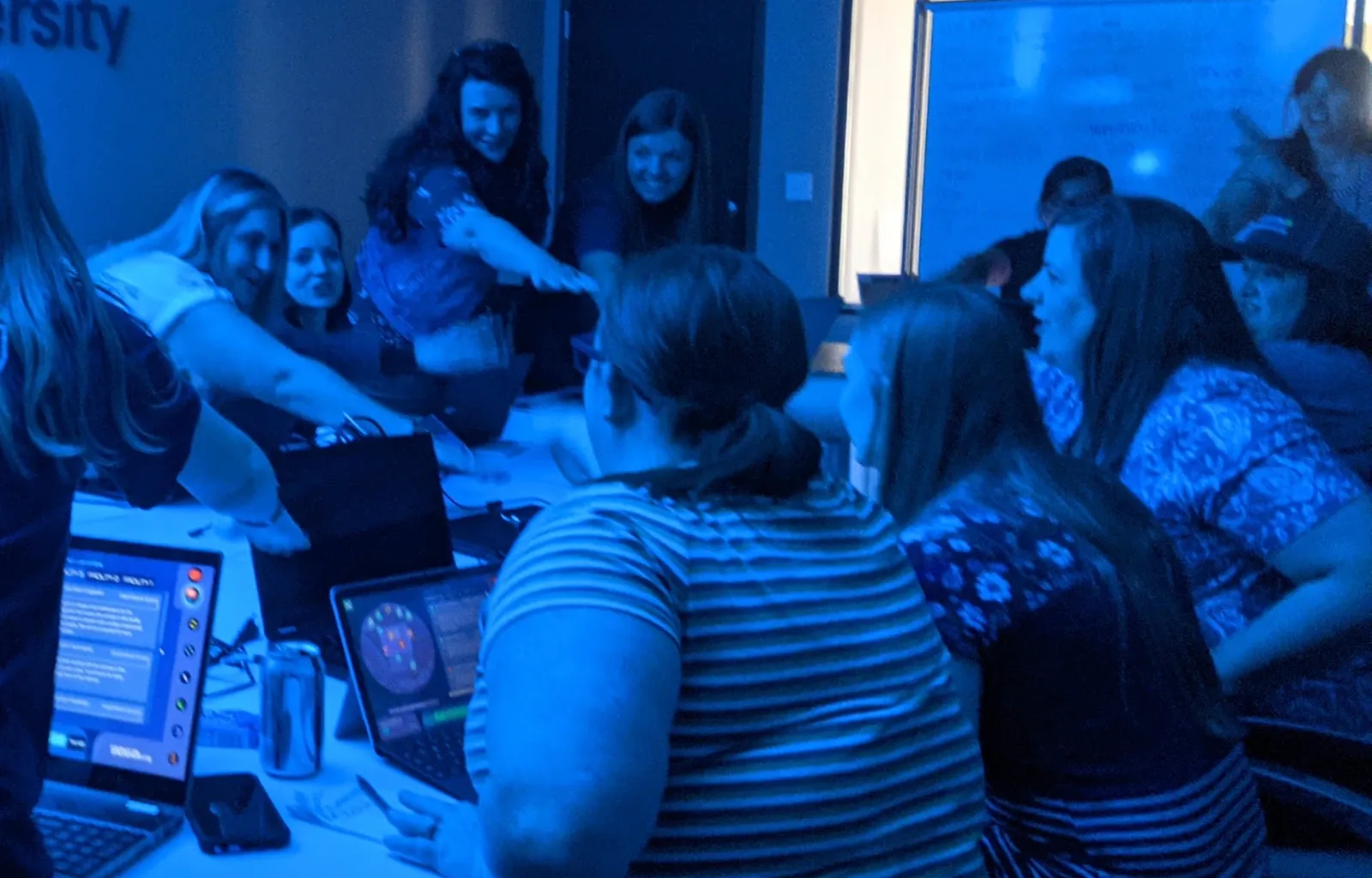 Teachers lean across a table toward one another, laughing over a decision, their laptops lit with the Mission.