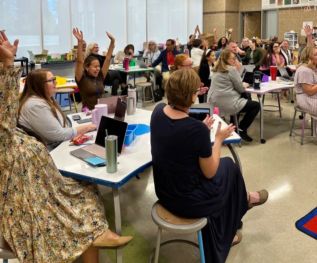 A full staff room of teachers at their laptops, several cheering with hands in the air.