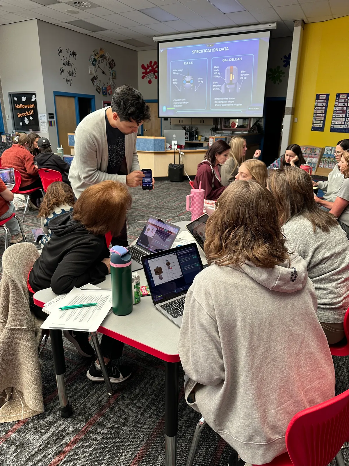 A facilitator leans in to help a table of teachers as the Mission’s data shows on the projector.