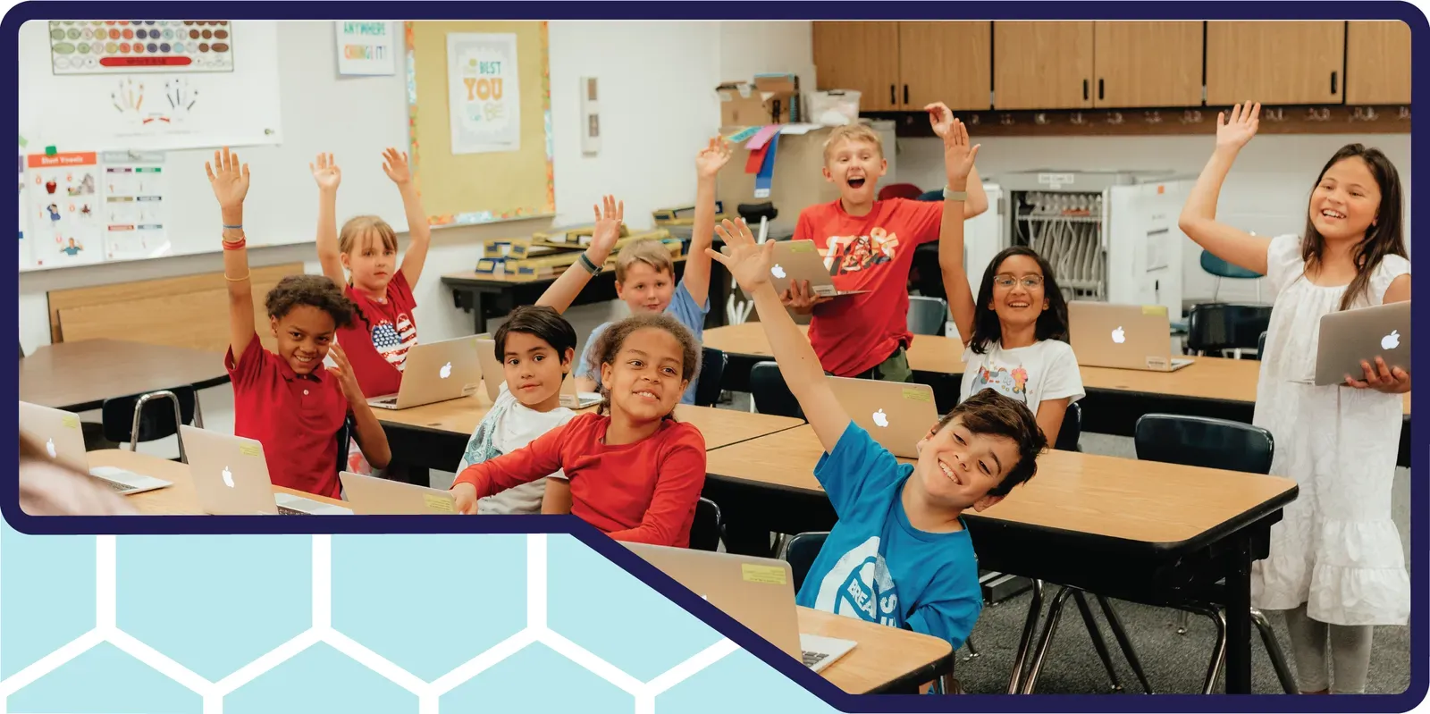 Smiling elementary students raise their hands at their desks with laptops in a bright classroom.