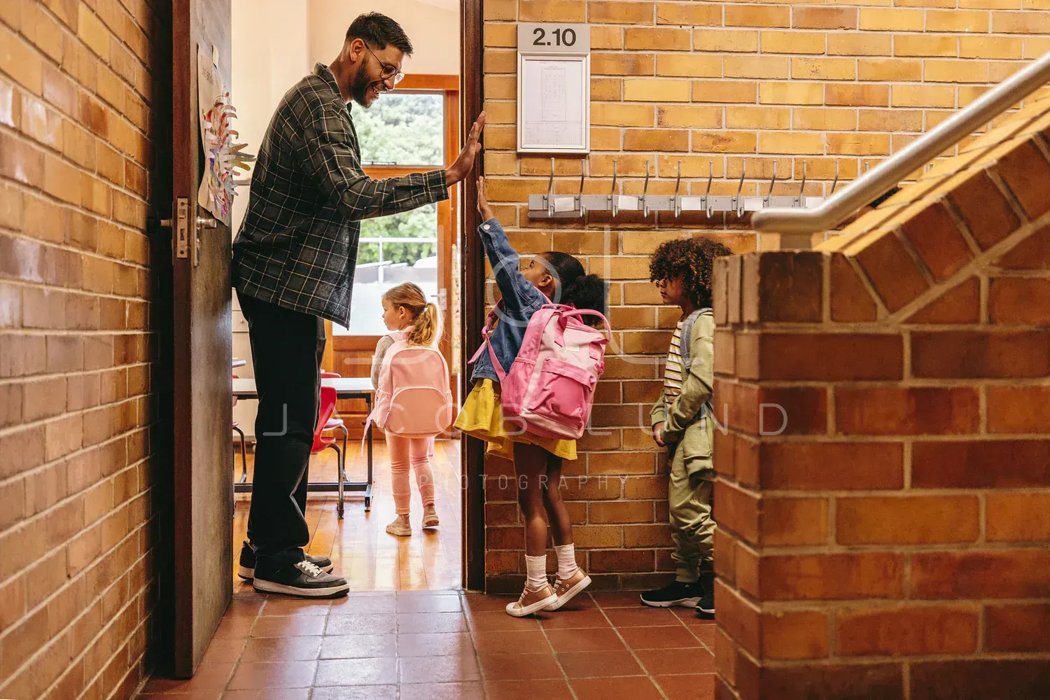 A smiling teacher at his classroom door high-fives a girl with a pink backpack as other students wait.