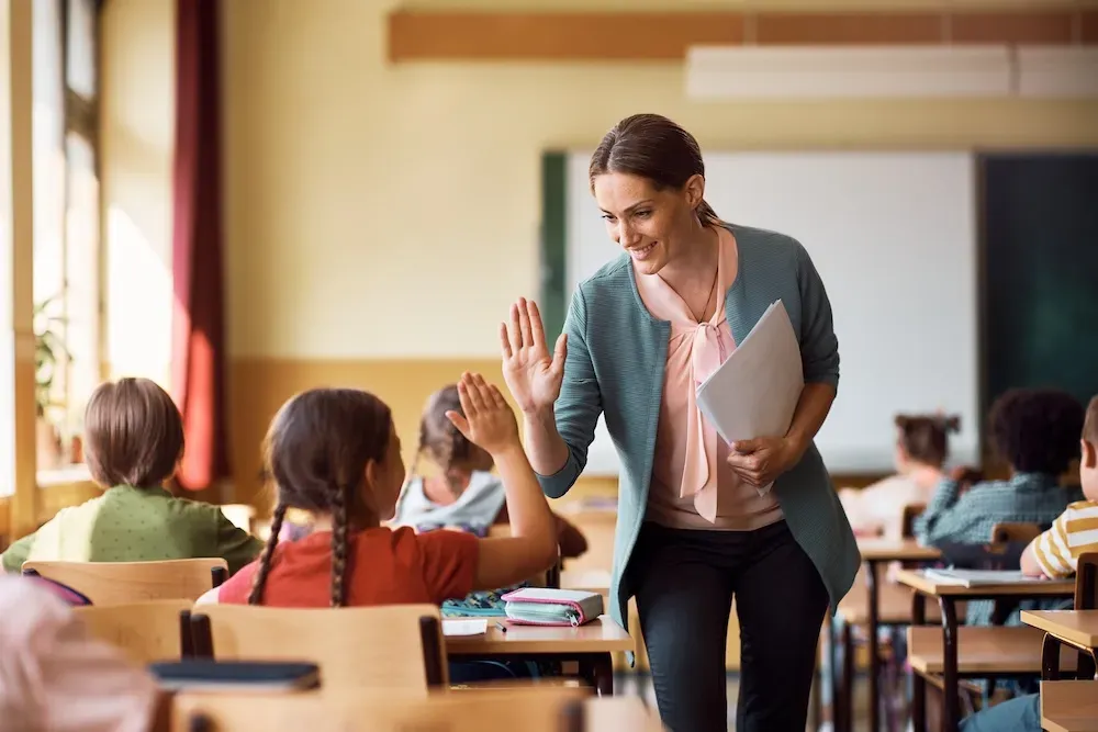 A smiling teacher high-fives a girl with braids at her desk in a sunlit classroom.